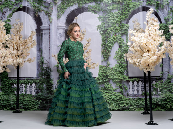 A woman wearing a modest ombre green tiered gown with a high neckline and long sleeves. The gown has a fitted bodice and a flowing skirt with multiple tiers of ruffles.