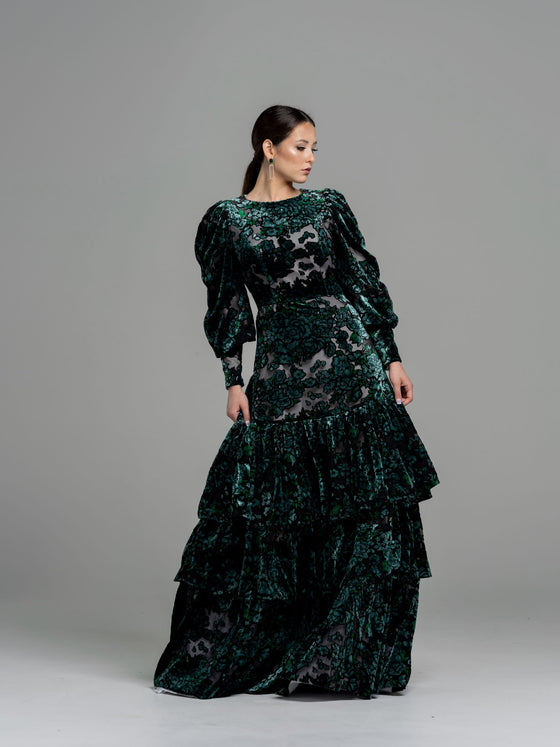 A woman wearing a modest dark green floral velvet burnout gown with puff sleeves. The gown has a fitted bodice, the bottom of the gown has 3 tiers of ruffles. 
