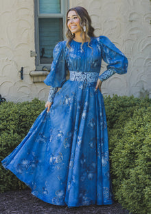  Woman wearing a dusty blue chiffon floral modest gown. The gown has volumnious puff sleeves and blue floral lace detailing at the waist and cuffs. 