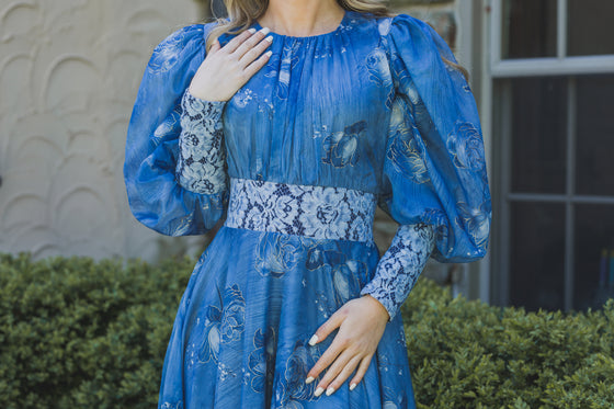 Woman wearing a dusty blue chiffon floral modest gown. The gown has volumnious puff sleeves and blue floral lace detailing at the waist and cuffs. 