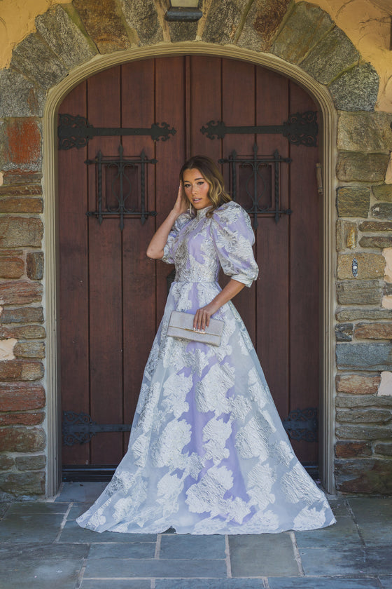 Woman wearing a modest lilac purple ballgown featuring a white and silver floral overaly with a high neckline and three quarter sleeve puff sleeves.