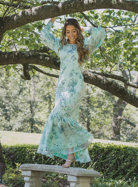 Woman wearing modest mint green gown with a floral print. The gown has full, voluminous sleeves and a ruffled bottom hem that adds a playful touch. 