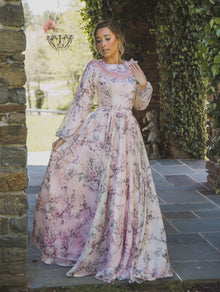  A woman wearing a modest blush pink gown with a floral print and ruffle collar detail. The floor sweeping gown has long sleeves. The floral print on the gown is soft and subtle, adding a touch of femininity
