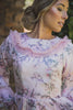 A woman wearing a modest blush pink gown with a floral print and ruffle collar detail. The floor sweeping gown has long sleeves. The floral print on the gown is soft and subtle, adding a touch of femininity