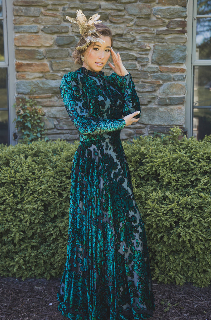 Woman wearing modest dark green floral velvet burnout gown with long sleeves that feature slight puff at the shoulders and a high collar neck. 