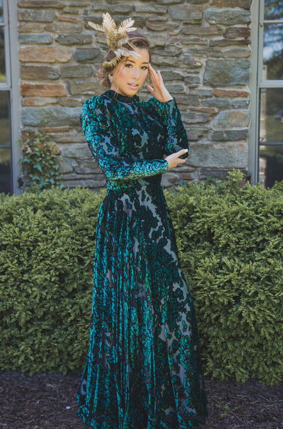 Woman wearing modest dark green floral velvet burnout gown with long sleeves that feature slight puff at the shoulders and a high collar neck. 