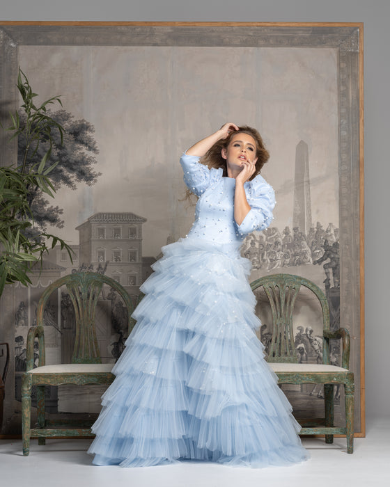 Woman wearing a modest powder blue gown with a beaded top and three-quarter length sleeves with a puff on top and straight cuffs. The gown has a tiered ball gown skirt. 