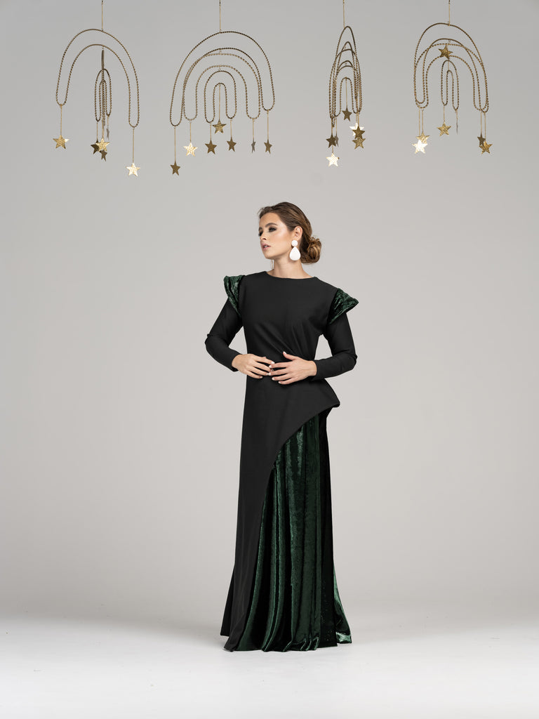 A woman wearing a modest green straight, long sleeve evening gown. The gown features a unique combination of green velvet on half of the skirt and green velvt shoulder detailing.