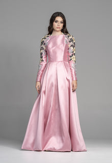  A woman wearing a modest pink box pleat gown with floral sleeves. The sleeves have raised embroidered florals and beading.