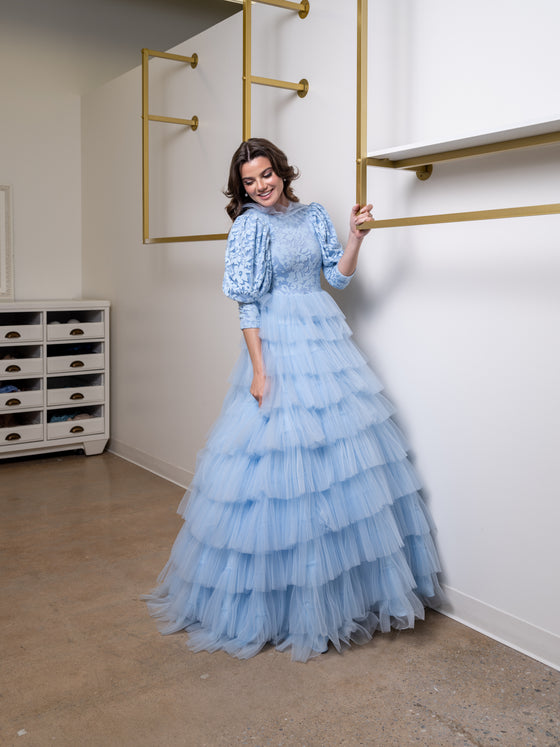 A woman wearing a modest powder blue ball gown. The skirt features layers of accordion tulle. The three-quarter voluminous sleeves are cut in a draped fabric allowing for maximum movement. The top is floral printed and has a high collar.
