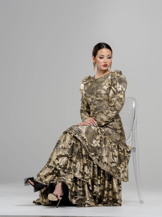 A woman sitting while wearing a modest gold floral pattern gown with voluminous sleeves. The gown has a fitted bodice and a flowing skirt with three tiers of ruffles. 