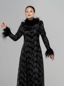  A woman wearing a black fitted modest gown with a high neckline and long sleeves. The gown has sequin detailing and a black feather collar and cuffs. 