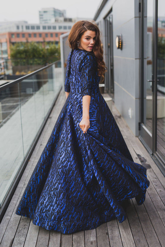 Woman wearing modest cobalt blue and black ball gown with cropped bolero. The three-quarter sleeve bolero has a large bow detail in front and mini covered buttons cascading down the back.