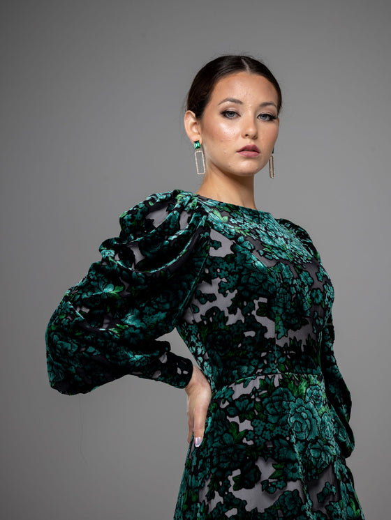 A woman wearing a modest dark green floral velvet burnout gown with puff sleeves. 