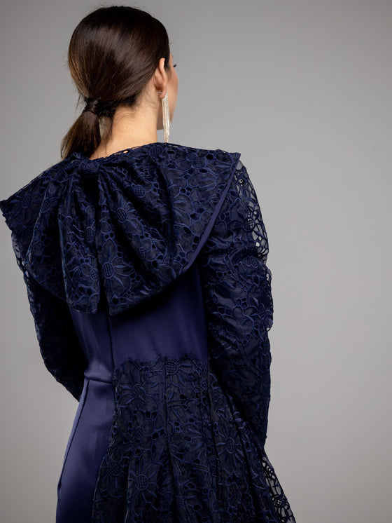 A woman wearing a modest blue navy lace gown with long sleeves. The gown has a large bow on top back.