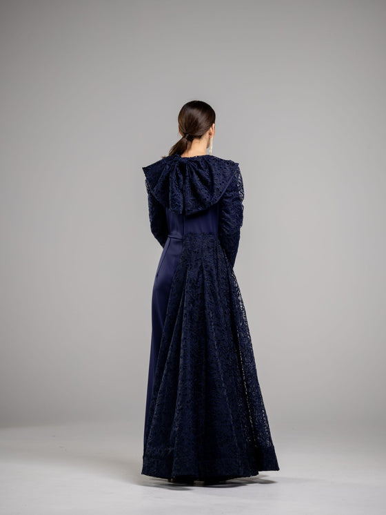 A woman wearing a modest blue navy lace gown with a high neckline and long sleeves. The gown has a large bow on top back.