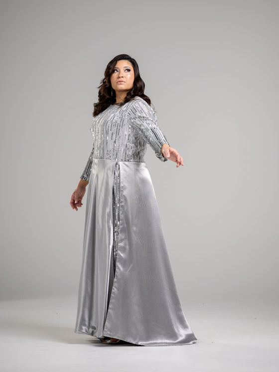 A woman wearing a silver modest straight silhouette gown adorned with sparkling sequins and a skirt cape. The gown has a high neckline and long sleees. 