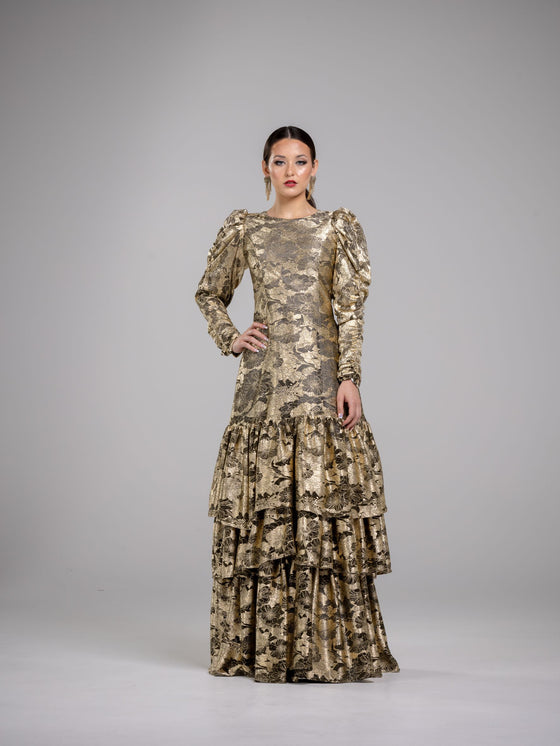 A woman wearing a modest gold floral pattern gown with voluminous sleeves. The gown has a fitted bodice and a flowing skirt with three tiers of ruffles. 