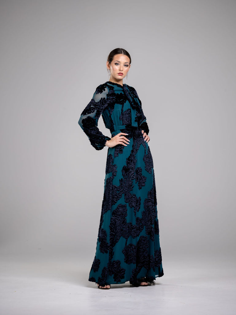 A woman wearing a modest dark green gown with velvet floral details. The gown has a matching tie neck detail tied in a bow. The velvet floral details are scattered across the bodice and sleeves of the dress, adding texture and depth to the overall design.