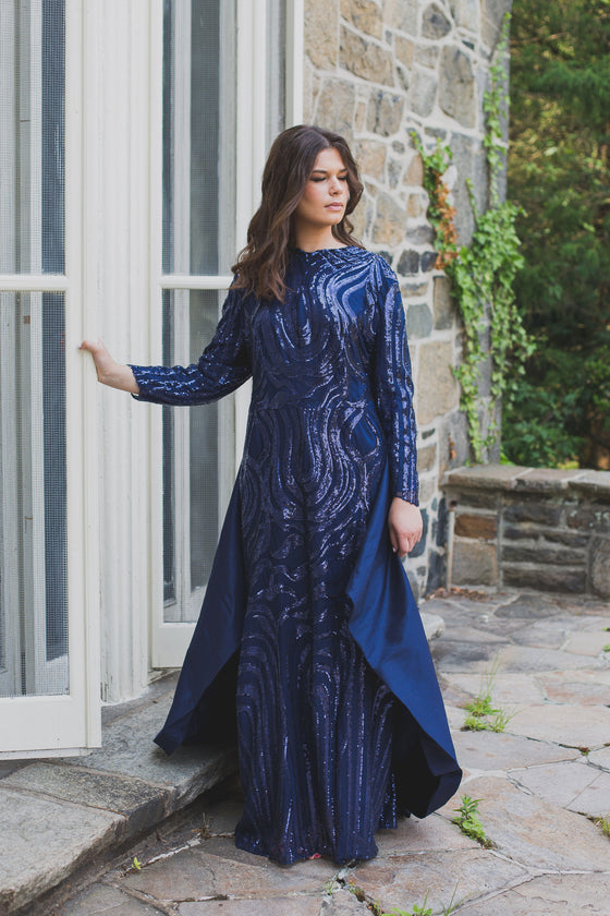 A woman wearing a modest navy blue gown with a shimmering sequin design and a removable skirt cape. The placement of the sequins is thoughtfully designed to compliment her curves.