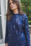 A woman wearing a modest navy blue gown with a shimmering sequin design and a removable skirt cape. The placement of the sequins is thoughtfully designed to compliment her curve