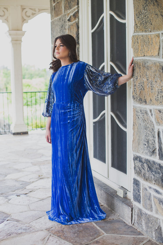 Woman wearing modest cobalt blue crushed velvet ruched gown with straight silhouette and billowy three-quarter length sleeves.