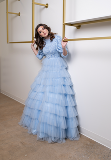  A woman wearing a modest powder blue ball gown. The skirt features layers of accordion tulle. The three-quarter  voluminous sleeves are cut in a draped fabric allowing for maximum movement. The top is floral printed and has a high collar.
