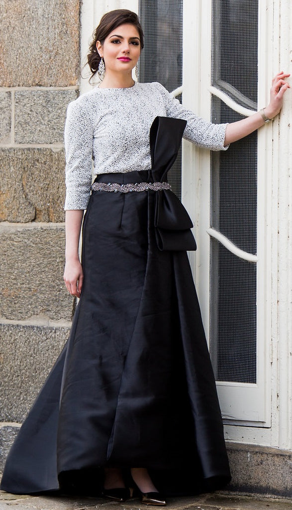 A woman wearing a modest black & white gown with a large bow by the waist.  The gown has a pearl Italian fabric top paired with a taffeta structured bold ballgown skirt with asymmetrical lines. The gown has three-quarter sleeves and a crew neck.