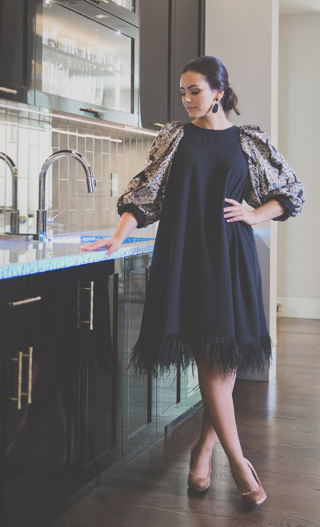 A woman wearing a modest midi length black cocktail dress. The dress has three-quarter sleeve bubble sleeves. The sleeves are adorned with a golden sequin printed abstract fabric, adding a touch of sparkle. The bottom of the dress features black feather detailing, adding a unique element. The dress is made from a lightweight crepe, making it comfortable and perfect for any occasion.