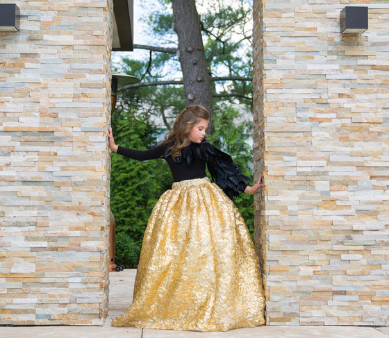 A girl wearing a modest black and gold two-piece ball gown with rich black feathers cascading down one sleeve and an imported gold crinkle foiled fabric ball gown skirt.