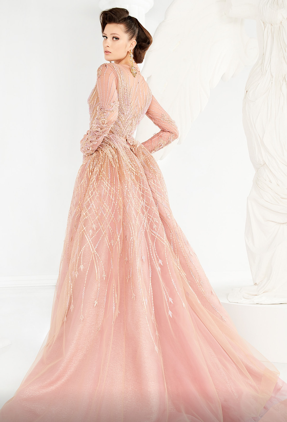 A woman wearing a pink, modest gown crusted with tonal beadwork of an intricate design. The embellished long sleeves add a touch of glamour, and the mermaid skirt gives the gown a sleek and sophisticated silhouette. The full-length gown is finished with a soft, open overskirt that flares from the waist to a court train, adding a touch of drama to the overall look.